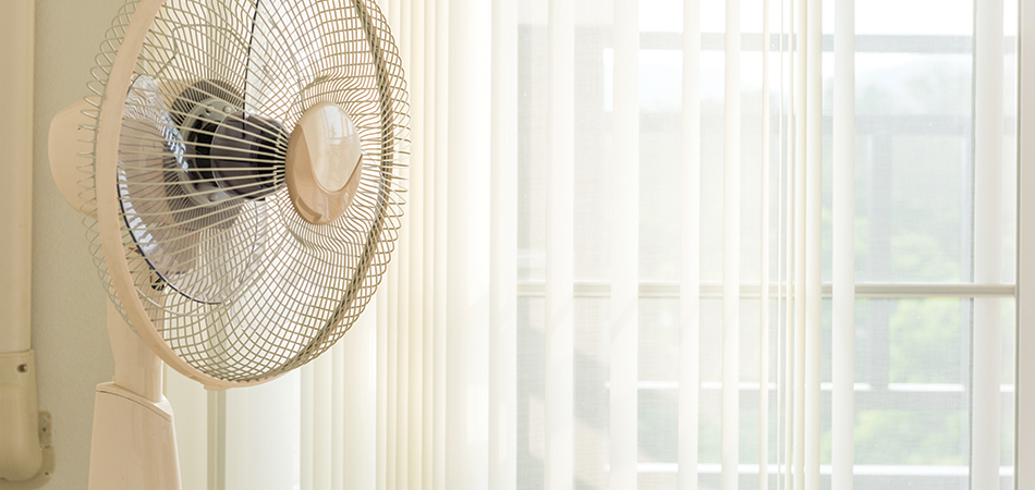 A white fan in front of a sunlit window that is covered with transparent yellow curtains.